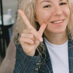 Crop happy adult female entrepreneur with blond hair in formal clothes smiling and showing index finger up sign while looking at camera sitting in armchair in cafe
